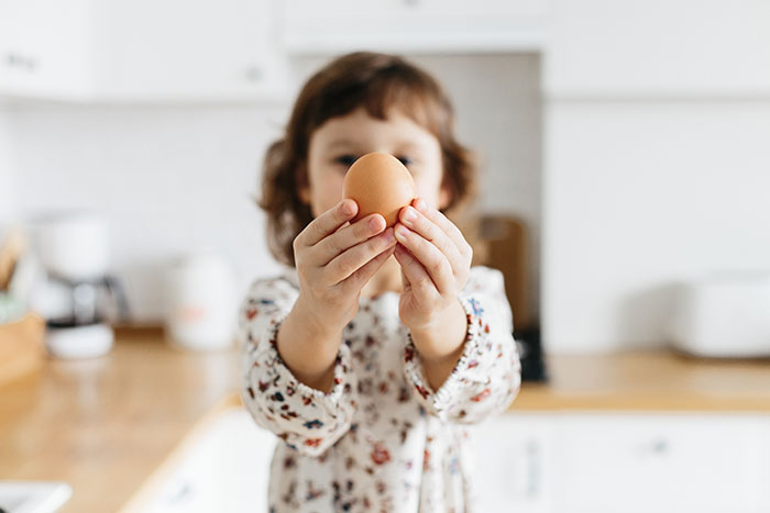 Child holding an egg in focus with blurred kitchen background, illustrating snooping parents discovering new sides of their kids.