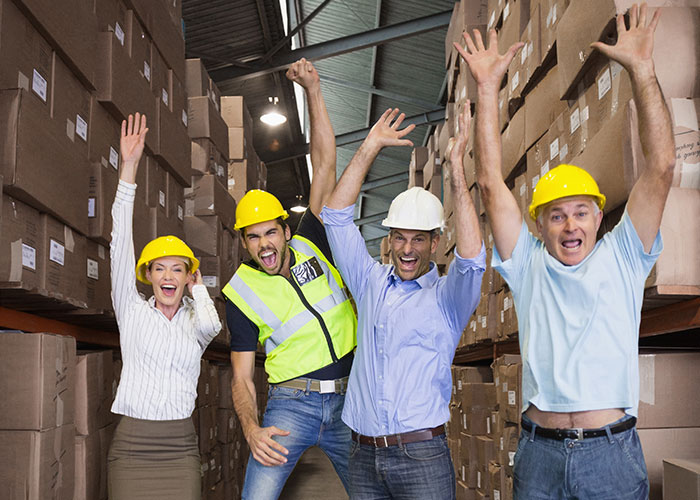 Four warehouse workers wearing helmets and safety gear celebrating with raised hands among stacked boxes at work.