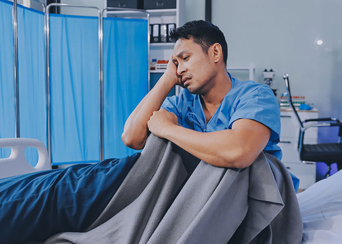 Healthcare worker sitting beside a hospital bed, looking stressed from the madness witnessed at work.