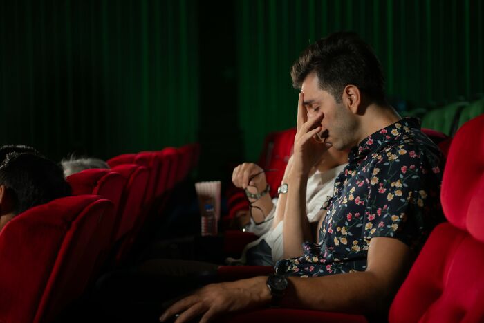 Man sitting in a theater with red seats covering his face, appearing rejected and distressed in a dim setting.