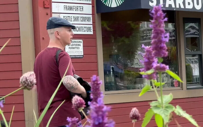 White man standing outside near Starbucks, visible tattoos and wearing a cap in daylight with purple flowers in foreground White man standing outside near Starbucks, visible tattoos and wearing a cap in daylight with purple flowers in foreground