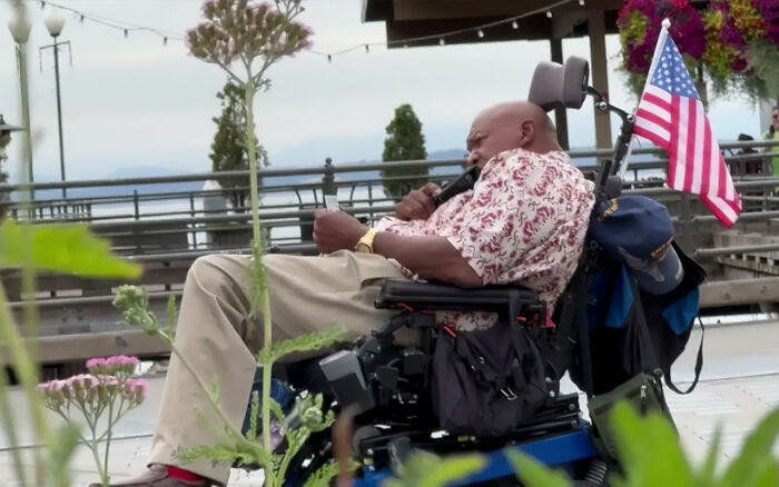 Black disabled veteran in a wheelchair outdoors with an American flag, holding a microphone near his mouth. Black disabled veteran in a wheelchair outdoors with an American flag, holding a microphone near his mouth.