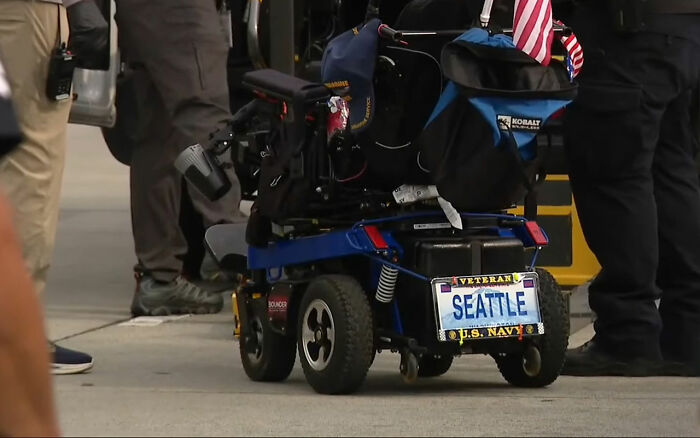 Power wheelchair with veteran license plate and U.S. Navy insignia, surrounded by people standing on city sidewalk. Power wheelchair with veteran license plate and U.S. Navy insignia, surrounded by people standing on city sidewalk.