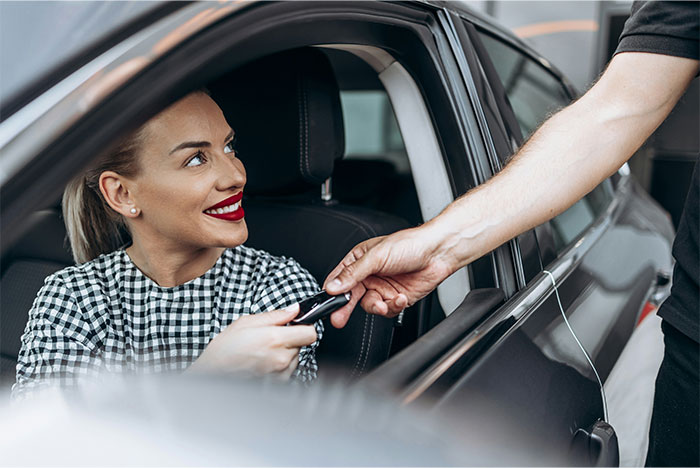 Woman sitting in a car smiling while handing car keys to a man outside at a dealership, showing a car purchase moment.