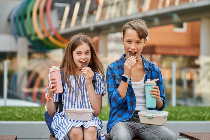 Two teens eating chocolate and holding drinks outdoors, illustrating the concept of being SAHM for teens and household chores debate.