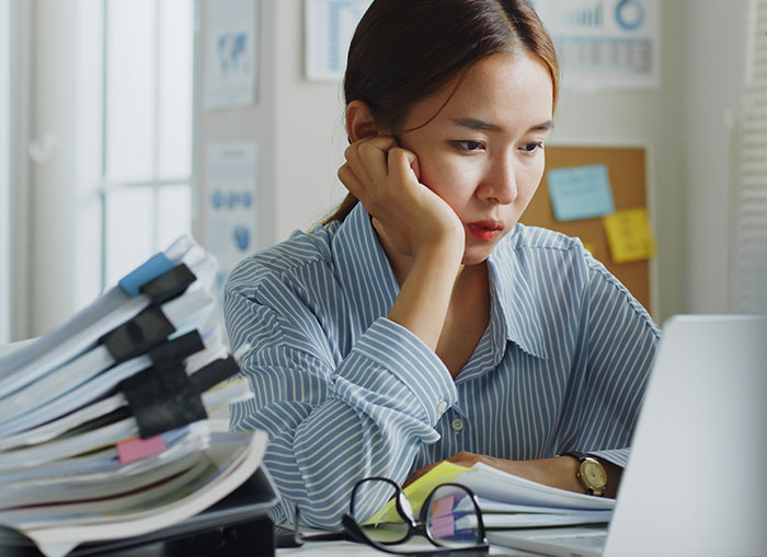 Young woman in striped shirt looking stressed at her laptop, surrounded by office files, reflecting workplace reprimand.