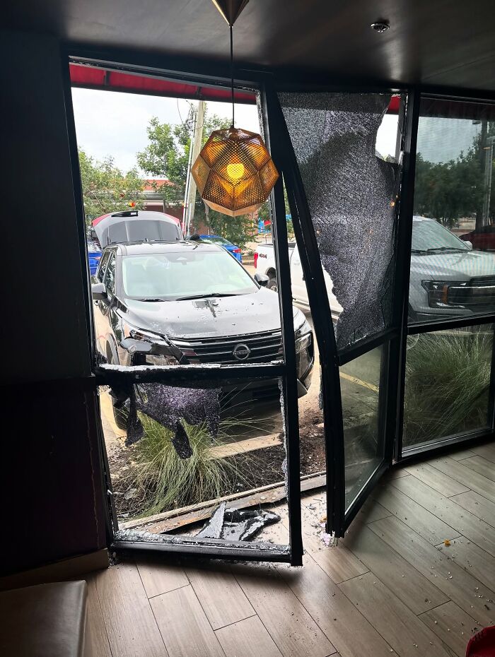 SUV crashed through shattered restaurant door with broken glass and damaged frame visible near parked vehicle outside.