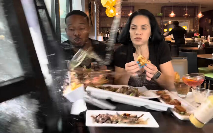 Couple eating inside restaurant as SUV crashes through window, causing glass and dishes to scatter violently.