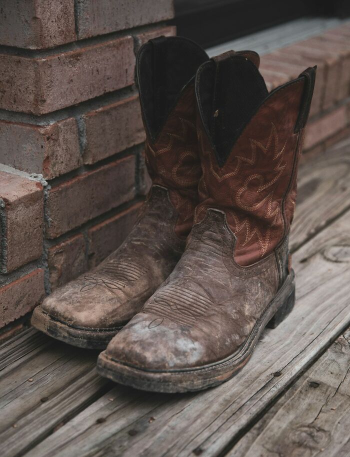 Worn cowboy boots resting on weathered wooden floor beside a brick wall, evoking eerie night shift workers atmosphere.