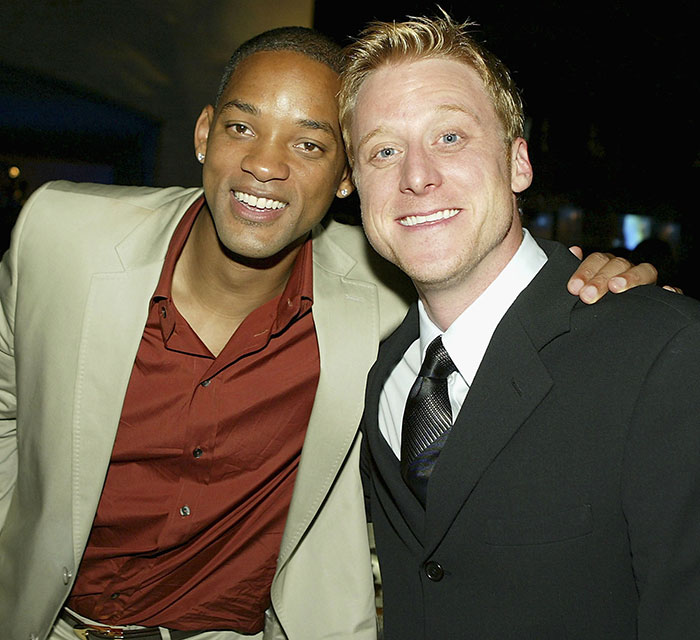 Will Smith's co-actor smiling with a man in a suit, highlighting studio name removal claim in publicity controversy.
