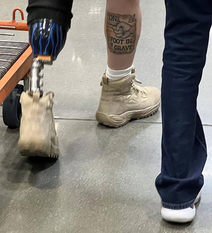 Person with prosthetic leg and tattoo reading "ONE FOOT IN THE GRAVE" on other leg, showing witty humor in public setting.