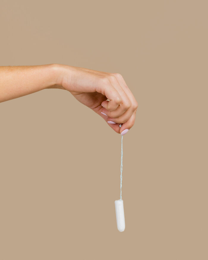 Hand holding a tampon by its string against a neutral background illustrating tampon left inside for a month story.