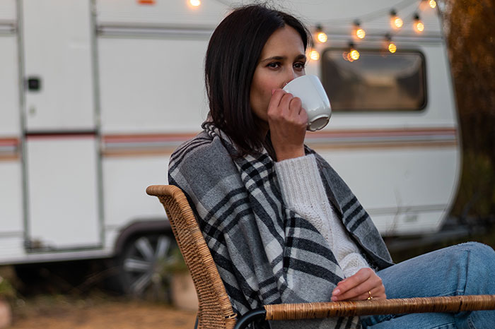 Woman regrets trip, sitting outside camper wrapped in blanket, sipping coffee with a pensive expression during tense travel moments.
