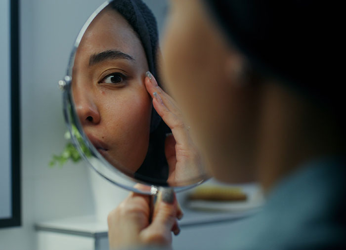 Woman examining her face in a handheld mirror, reflecting concerns related to the Love Island face trend and filler blindness.