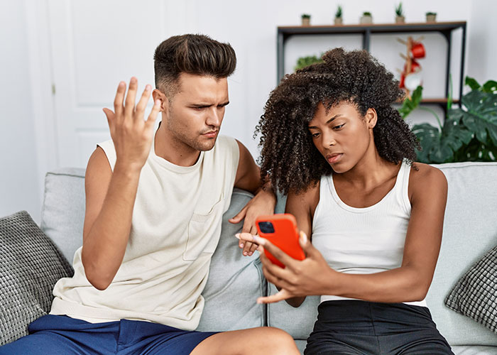 Woman checking boyfriend&rsquo;s phone with a worried expression after a jealous freak-out while sitting on a couch together.