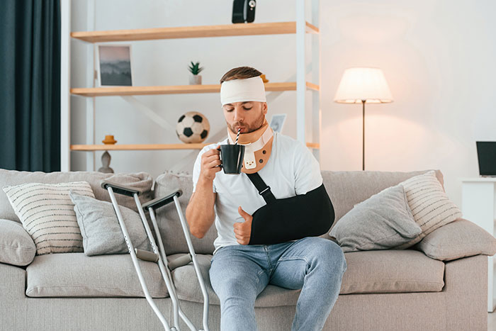 Injured boyfriend with arm sling and head bandage sitting on couch, drinking from cup, with crutches nearby at home.