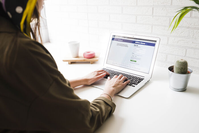 Person typing on a laptop at a white desk, browsing a social platform, surrounded by a cactus and a donut on a stand.