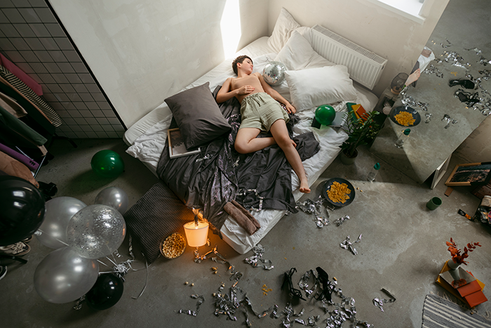 Young man lying amid party mess and scattered balloons in a bedroom showing aftermath of temporary houseguest chaos. Young man lying amid party mess and scattered balloons in a bedroom showing aftermath of temporary houseguest chaos.