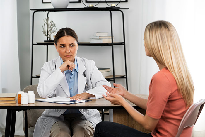 Woman in a medical consultation expressing chronic pain, doctor listening skeptically in a clinical setting.