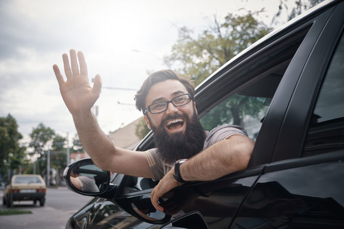 Man with glasses and beard happily waving from the driver’s seat of a black car, outside on a bright day. - 21
