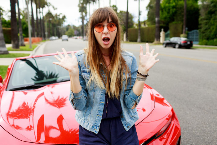 Woman in sunglasses and denim jacket reacting with surprise in front of a red car, illustrating lady in brand new Lexus scenario. - 1