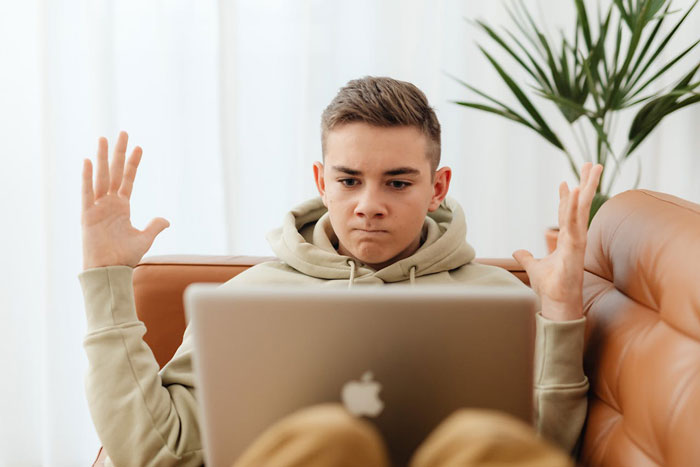 Teen boy sitting on couch with raised hands, looking frustrated at a laptop, related to autistic nephew babysitting issue.
