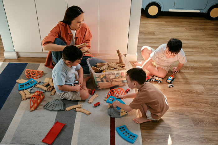 Woman babysitter playing with three children on floor, surrounded by wooden train tracks and toy vehicles in a home setting.