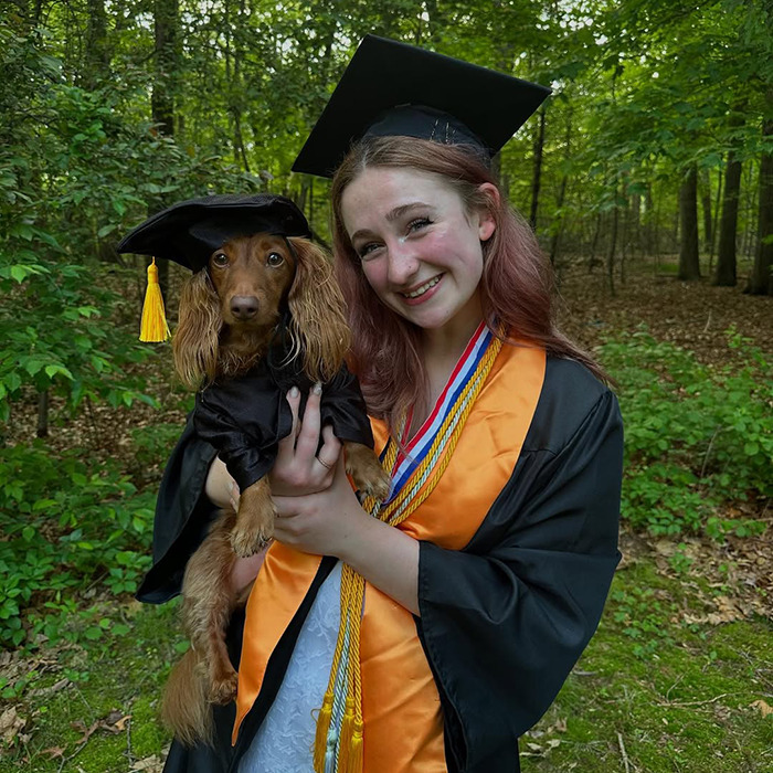 Young woman joining anti-loneliness club celebrates graduation outdoors holding a dog also dressed in graduation attire.