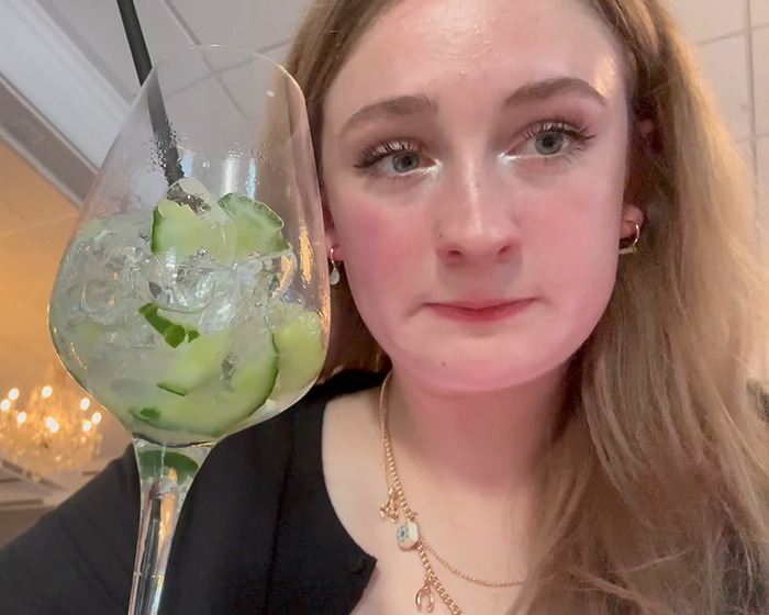 Young woman sitting alone in a restaurant, holding a glass with cucumber and ice, joining anti-loneliness club.