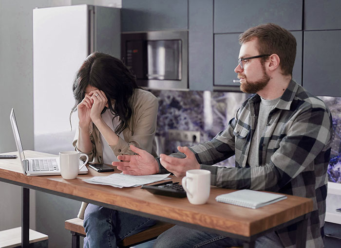 Woman feels financially trapped as jobless fianc&eacute; argues at kitchen table with bills and laptop nearby.