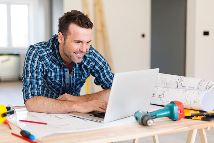 Man in plaid shirt working on laptop surrounded by tools and blueprints, representing financially trapped woman scenario.