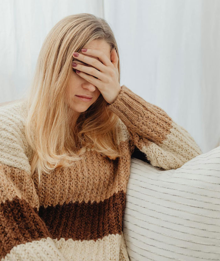 Woman in a cozy sweater sitting on a couch with hand covering her face, reflecting postpartum trip and family conflict.