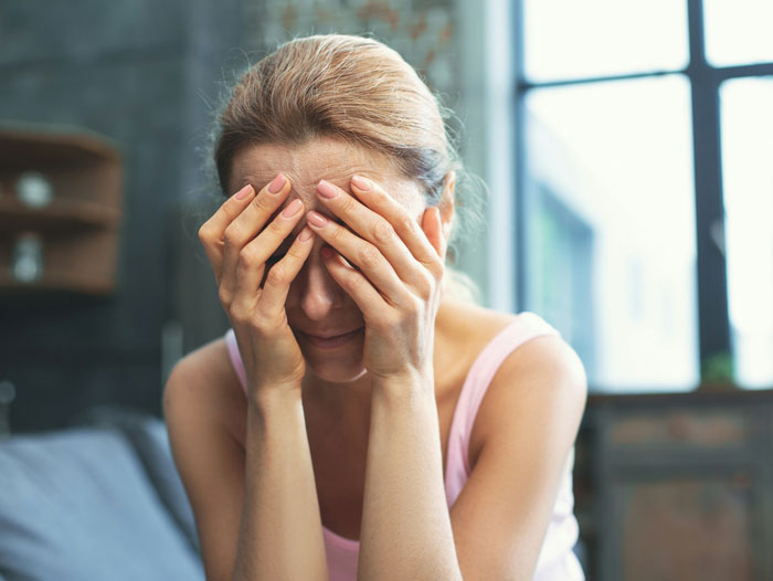 Stressed woman sitting indoors with hands covering her face, reflecting postpartum trip and family tension.