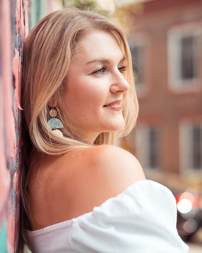 Young woman wearing a smart ring, smiling thoughtfully while leaning against a colorful wall outdoors.