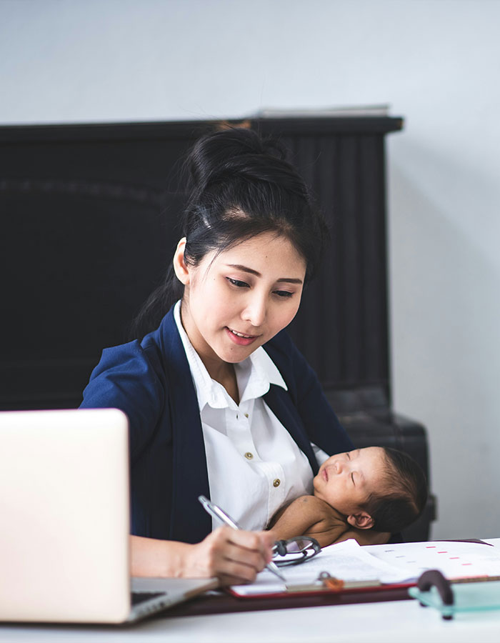 Working mom in the U.S. multitasking with baby in arms while writing notes at her desk with a laptop and documents. Working mom in the U.S. multitasking with baby in arms while writing notes at her desk with a laptop and documents.