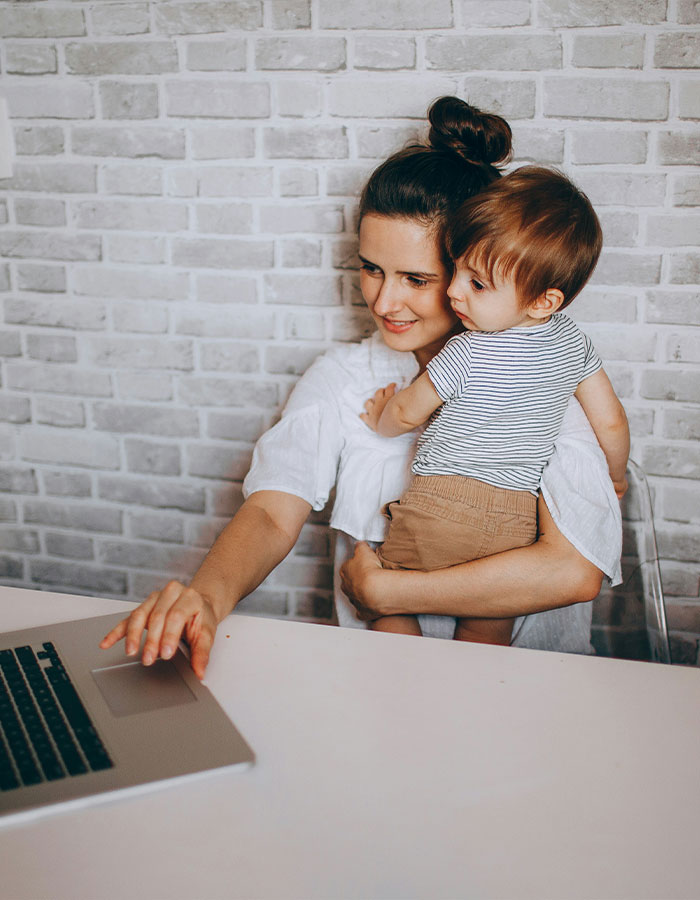 Working mom in the U.S. multitasking with child in her arms while using a laptop at home against a brick wall background. Working mom in the U.S. multitasking with child in her arms while using a laptop at home against a brick wall background.