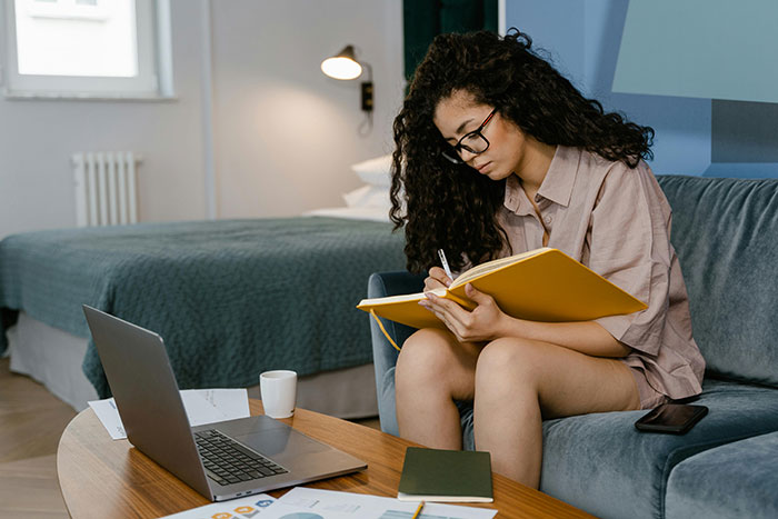 Young woman writing in a notebook at home, surrounded by laptop and papers, illustrating college application lies theme.