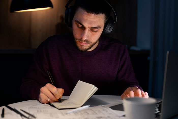 Student wearing headphones writing in notebook late at night with papers and laptop, illustrating college application lies.
