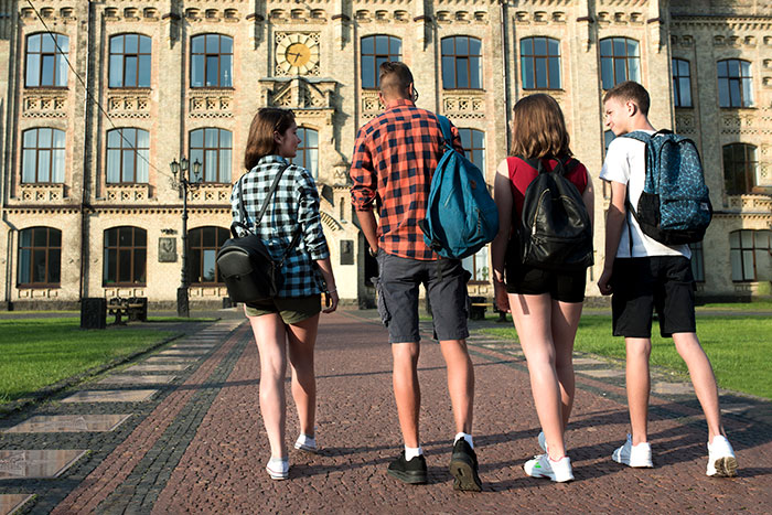 Four students with backpacks walking toward college building on a sunny day representing college application challenges and lies.