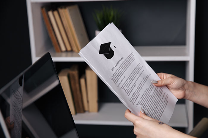 Person holding college application papers with graduation cap icon near a laptop and bookshelf in background.