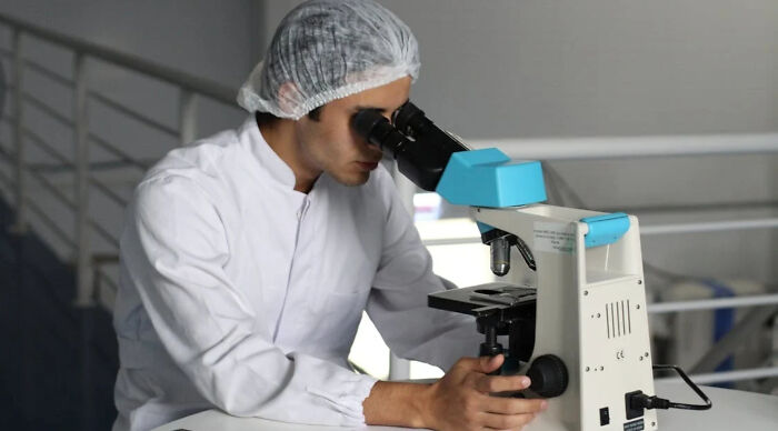 Scientist in lab coat and hairnet using microscope, focused on research in a modern laboratory setting for Would You Rather Poll 3.