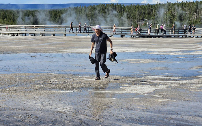 Visitor walking across forbidden area in Yellowstone National Park while others stay on boardwalk with steam rising nearby. - 3