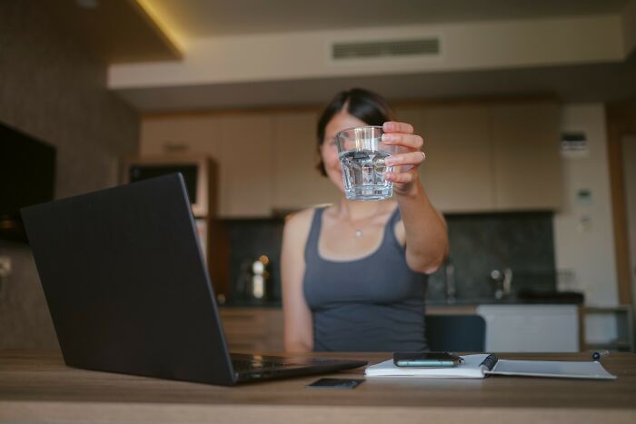 Woman holding a glass of water in front of a laptop, demonstrating hydration as a key tip for how to lose weight.