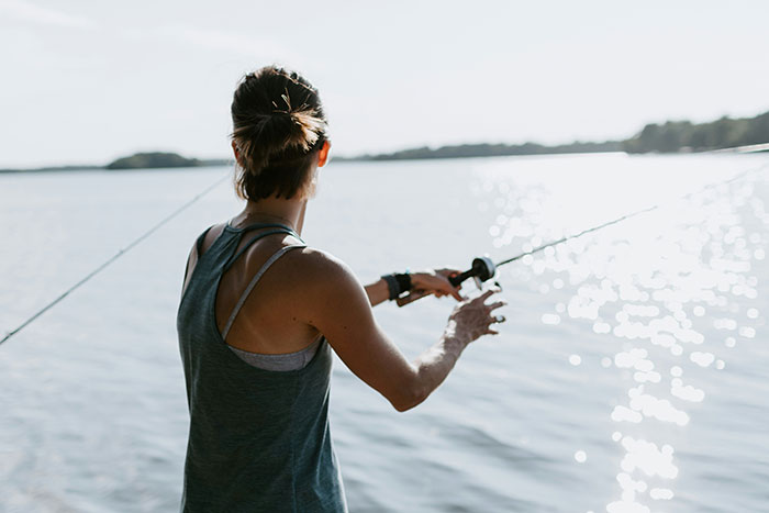 Young girl fishing outdoors by a lake, holding a fishing pole with the sun reflecting on the water surface.