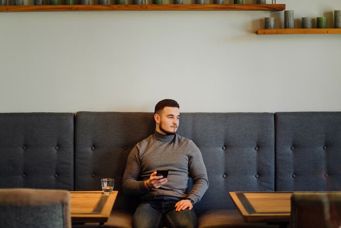 Young man sitting alone on a couch, holding a phone and looking away, illustrating stories of people getting savagely rejected.