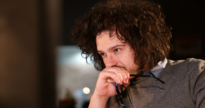 Young man with curly hair holding glasses, deep in thought, illustrating concepts of words that don’t exist in English.