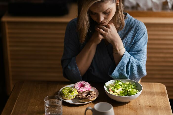 Woman in a blue robe contemplating food choices with a salad and donuts on the table, focused on weight loss tips.