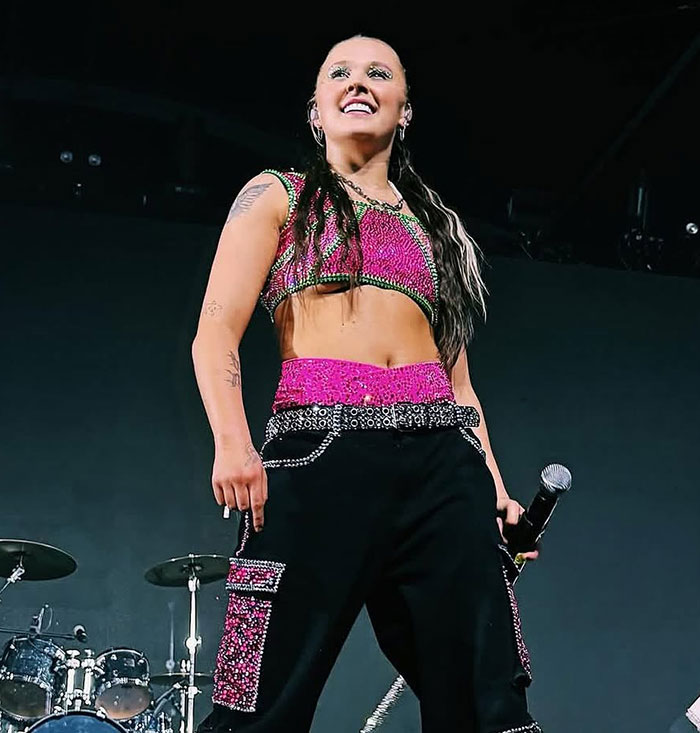 JoJo Siwa smiling on stage wearing a pink and black outfit during a live performance with microphone in hand.
