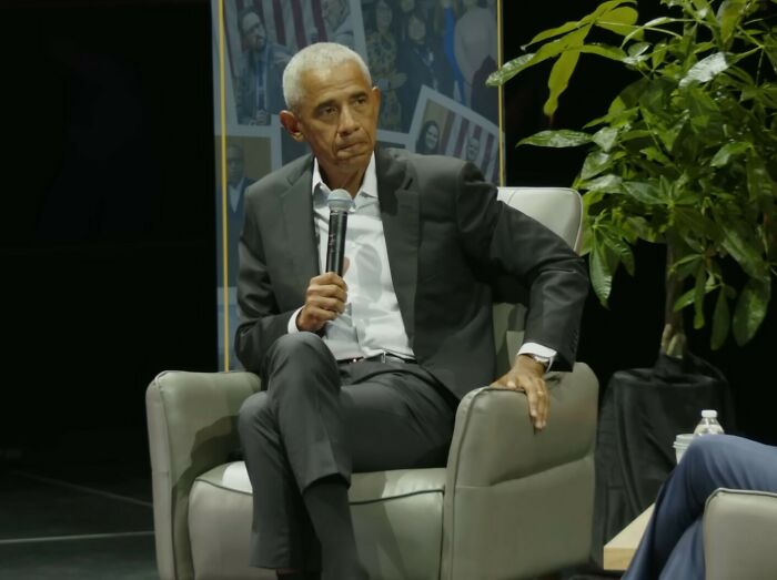 Barack Obama seated in armchair holding microphone, discussing post-presidency challenges with wife Michelle on stage. Barack Obama seated in armchair holding microphone, discussing post-presidency challenges with wife Michelle on stage.