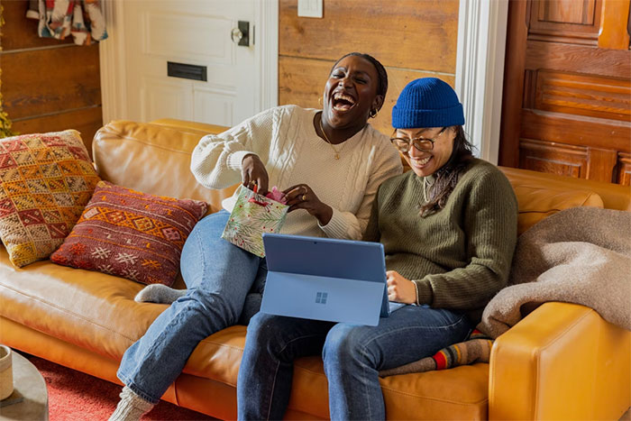 Two women sharing a laugh on a couch, one holding a gift bag while they look at a laptop together.